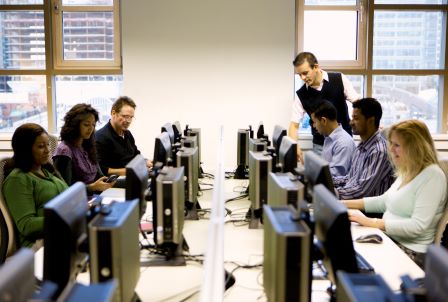 ICDL Test Center Beispielbild, Menschen die vor Computern sitzen und eine stehende Person, die sich über Computer beugt